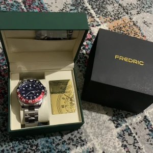 Fredric Dual Time sportklocka stål - Snygg sportklocka från Fredric med dual time-funktion. Klockan har en rund blå urtavla, röd och blå bezel med vita siffror, och ett robust armband i blankt rostfritt stål. Levereras i originalask med certifikat. Perfekt för dig som gillar stil och funktion.