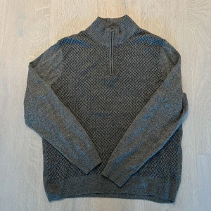 Grå stickad tröja med half zip från Dockers - Snygg grå stickad tröja från Dockers i storlek M. Tröjan har ett diskret mönster framtill, ribbad krage och en praktisk half zip. Långärmad och tillverkad i 100% akryl, perfekt för kyliga dagar. Enkel att matcha med jeans eller chinos.