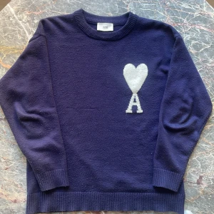 Mörkblå stickad tröja från Ami - Snygg mörkblå stickad tröja från Ami med ett stort vitt hjärta och bokstaven A på bröstet. Tröjan har rund hals, ribbade muddar och lång ärm. Perfekt för dig som gillar stilrena detaljer och vill ha något som sticker ut lite extra.