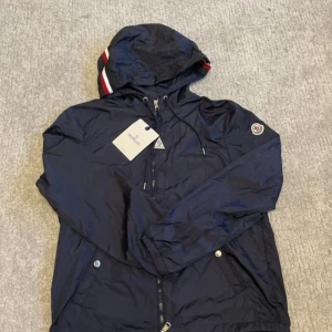 Mörkblå vindjacka från Moncler - Snygg mörkblå vindjacka från Moncler med huva och coola rödvita detaljer på kanten. Jackan har dragkedja framtill, snörning vid huvan och Moncler-logga på ärmen. Materialet är lätt och syntetiskt, perfekt för blåsiga dagar. Fickor med tryckknappar och klassisk Moncler patch på insidan. Taggarna är ej kvar på jackan 