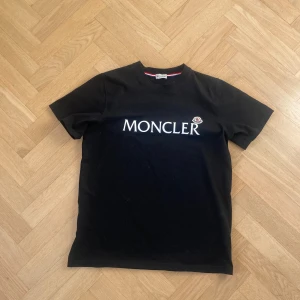 Svart Moncler t-shirt med logga - Svart t-shirt från Moncler med stor vit logga tryckt över bröstet och klassisk rund hals. T-shirten har korta ärmar och är tillverkad i mjuk bomull. Perfekt för en clean och stilren look.