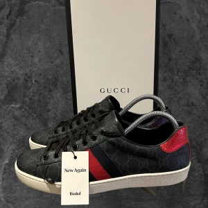 Svarta gucci ace skor  - Gucci ace sneakers i svart med diskret GG-monogram, klassisk röd och marinblå rand på sidan samt kontrasterande röd och mörkblå häl i ormskinnsmönster. Platt vit sula med unikt mönster undertill och snörning framtill. Snygg och lyxig design som sticker ut.