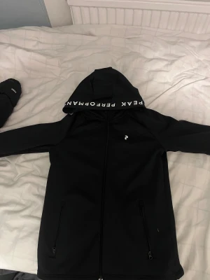   Peak Performance tränings kofta - Svart tränings kofta från Peak Performance med huva och vit logga på bröstet. Samt text på luvan. Perfekt för träning eller att ha som en vanlig tröja.