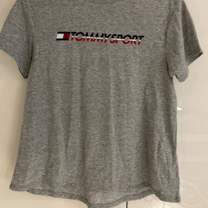 Grå t-shirt Tommy Sport - Snygg grå t-shirt från Tommy Sport med tryckt logga i svart, rött och vitt på bröstet. Klassisk rund hals och korta ärmar. Perfekt för dig som gillar sportig och clean stil. Materialet är mjuk bomull som känns skönt mot huden.