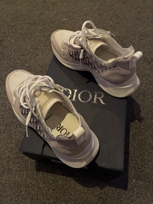  sneakers med monogram och mocka - Snygga sneakers i vit och beige med klassiskt monogram på sidan. Skorna har rund tå, platt sula och snörning. Materialmix med mesh och mocka ger en lyxig vibe. Perfekta för dig som vill sticka ut med designer på fötterna.