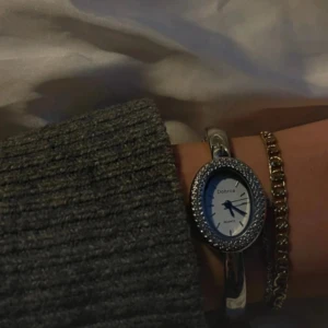 Vintage Silvrig oval klocka  - Snygg silvrig klocka  med oval urtavla och glittrande stenar runt ramen. Klockan har ett stilrent armband i metall och en vit urtavla med svarta detaljer. Perfekt accessoar för att lyfta din outfit. Batteridriven 🩷 kom privat för frågor 💞