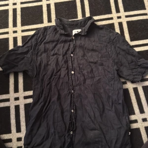 Svart linneskjorta från NN07, M - Svart skjorta från NN07 i storlek M, tillverkad i mjukt linne. Klassisk krage, korta ärmar och knappstängning framtill. En bröstficka på vänster sida ger en chill vibe. Perfekt för dig som gillar stilren och enkel design.