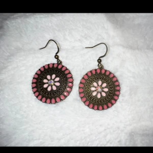 Rosa blommiga örhängen i boho-stil - Säljer ett par runda örhängen med antikguldfärgad metall och detaljer i rosa och ljusrosa. Örhängena har ett blommönster i mitten och en kant med rosa droppformade detaljer. Perfekta för dig som gillar boho och färg!