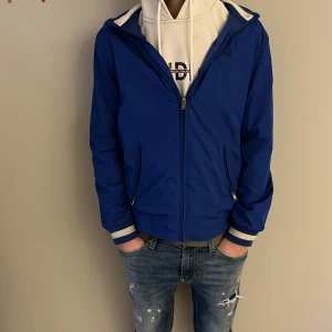 Blå vindjacka från Polo Ralph Lauren - Snygg blå vindjacka från Polo Ralph Lauren med huva och vita detaljer på muddar. Jackan har dragkedja framtill, två fickor med zip och en stilren design. Perfekt för dig som gillar clean stil. Huvan går att lägga in i jackan så man kan välja att ha luvan på eller av 