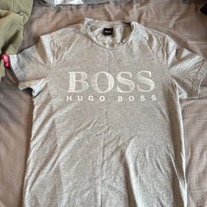 Grå t-shirt från Hugo Boss - Snygg grå t-shirt från Hugo Boss med stort vitt BOSS-tryck på bröstet. Tillverkad i mjuk bomull och har klassisk rund hals. Perfekt basic-plagg med stilren design och bekväm passform.