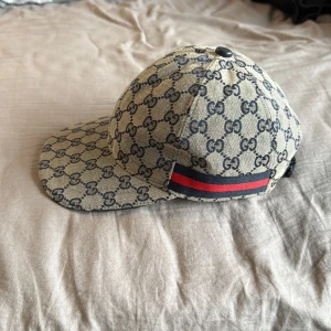 Gucci beige keps med GG-mönster - Snygg beige keps från Gucci med klassiskt GG-mönster i mörkblått över hela kepsen. På sidan finns en ikonisk röd och marinblå rand. Justerbar rem baktill i svart skinn.