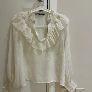 Vit blus med volang från Zara - Supersöt vit blus från Zara med volang längs v-ringningen och ärmar med breda smockade muddar. Blusen har diskret prickigt mönster och är gjord i ett lätt, luftigt material som ger en romantisk vibe. Perfekt för dig som gillar detaljer och en feminin stil.