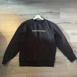 Svart Peak Performance hoodie storlek M  - Svart hoodie från Peak Performance i storlek M med stort vitt logotryck på bröstet. Klassisk rund hals och ribbade muddar vid ärmslut och nederkant. Perfekt för chill dagar och enkel att matcha med jeans och mjukisbyxor 