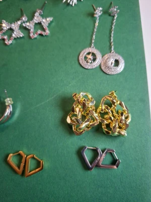 Mix av örhängen i guld och silver - Säljer ett gäng coola örhängen i olika former och färger – bland annat stora blommor i guld, chunky hoops i silver, stjärnformade örhängen med glittriga stenar, samt örhängen med blommor och fransar. Perfekt för dig som vill variera din stil och sticka ut.
