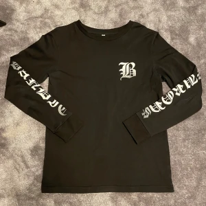 Bandit europe long sleeve - Svart långärmad tröja från Bandit europe med stort silvrigt B-tryck på bröstet och ryggen. Ärmarna har coolt silvrigt texttryck i gotisk stil. Mjuk och skön bomull, perfekt för dig som gillar streetwear och statement-plagg.