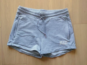 Grå Better Bodies shorts med snörning, stl M, dam - Snygga grå shorts från Better Bodies i mjuk bomullsmix. De har elastisk midja med snörning, två sidofickor och vit logga på benet. Passformen är normal och modellen är midwaist, perfekt för träning eller chill. Klassisk stil med enkel design.