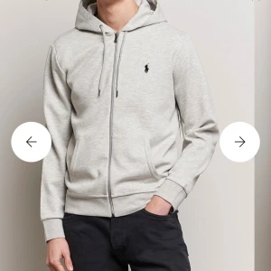 Ljusgrå hoodie från Polo Ralph Lauren - Snygg ljusgrå hoodie från Polo Ralph Lauren med dragkedja, huva och snörning. Tröjan har två fickor fram och den klassiska svarta Polo-loggan på bröstet. Perfekt för chill dagar och enkel att matcha med jeans eller joggers.