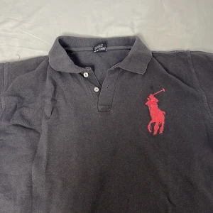 Svart/grå  piké från Ralph Lauren L - Svart pikétröja från Ralph Lauren med stor röd broderad logga på bröstet och röd siffra 3 på ärmen. Klassisk krage, knappar framtill och korta ärmar. Tillverkad i bomull för en skön känsla.