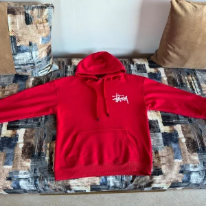 Röd Stüssy hoodie med logga - Säljer en röd hoodie från Stüssy i storlek M. Tröjan har stor vit Stüssy-logga på ryggen och mindre logga på bröstet. Klassisk huva med snörning och magficka framtill. Materialet är mjuk bomull och passformen är normal.