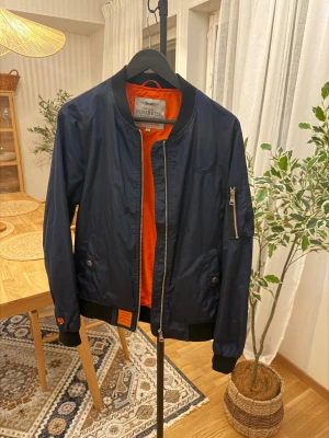 Mörkblå bomberjacka från Bombers - Snygg mörkblå bomberjacka från Bombers med orange foder, klassisk ribbad krage och muddar. Jackan har dragkedja framtill, fickor med tryckknappar och en extra ficka med dragkedja på ärmen. Perfekt för dig som gillar streetstyle.
