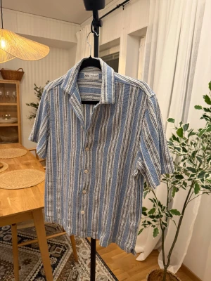 Randig blå skjorta från Zara - Snygg kortärmad skjorta från Zara med blå och vita ränder. Skjortan har klassisk krage, knappar framtill och är tillverkad i ett mjukt bomullsmaterial som känns chill och somrigt. Perfekt för dig som gillar avslappnad stil med lite retrovibe.
