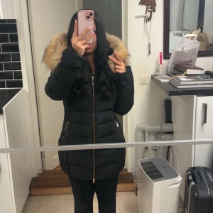 Svart Hollies dunjacka med päls - Säljer en svart dunjacka från Hollies med fluffig päls på huvan och snygga guldfärgade dragkedjor. Jackan är quiltad och har lång ärm, perfekt för kalla dagar. Knappt använd, priset går även att diskuteras🥰.