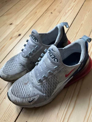 Nike Air Max 270 vita och röda sneakers - Nike Air Max 270 sneakers i vitt med röda och marinblå detaljer. Skorna har en synlig röd Air Max-sula, mesh-överdel med ventilerande hål och snörning. Swoosh-logga i rött och svart på sidan och fram. Snygg sportig look med platt sula och rund tå.