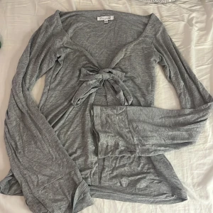 Grå långärmad knytblus Design by Si - Supersnygg grå långärmad blus från Design by Si i mjuk bomullsmix. Djup v-ringning och knyt framtill. Perfekt till jeans eller kjol. Modellen är croppad och har en loose passform.