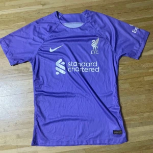 Lila Liverpool FC Nike fotbollströja - Säljer en lila Liverpool FC fotbollströja från Nike med korta ärmar. Tröjan har klubbens logga och sponsortryck på bröstet samt tryck på båda ärmarna. Materialet är lätt och andas, perfekt för träning eller match. Snygg och sportig design för alla Liverpool-fans.