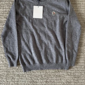 Grå stickad tröja från Moncler - Snygg grå stickad tröja från Moncler med klassisk rund halsringning och broderad logotyp på bröstet. Tröjan har ribbade muddar vid ärmslut och nederkant, perfekt för lager-på-lager under kyliga dagar.