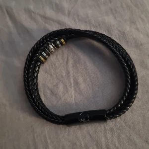 Svart flätat armband med detaljer - Coolt svart armband med dubbla flätade band och metallspänne. Armbandet har flera ringformade detaljer i silver, guld och svart, varav en med döskalle. Perfekt accessoar för dig som gillar edgy stil och snygga detaljer.