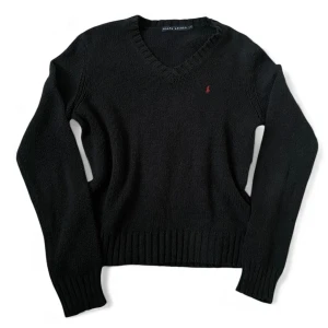 Svart stickad tröja från Ralph Lauren | 40  - Svart stickad tröja från Ralph Lauren med v-ringning och diskret röd logga broderad på bröstet. Tröjan har långa ärmar, ribbade muddar och en klassisk passform som funkar till allt. Perfekt för dig som gillar stilrena och tidlösa plagg.