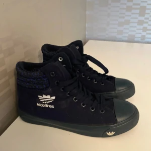 Mörkblå höga sneakers från Midellinss - Säljer ett par mörkblå höga sneakers från Midellinss med snörning och vit logga på sidan. Skorna har en rund tå, platt sula och detaljer i textil runt ankeln. Perfekta för dig som gillar en sportig och urban stil.