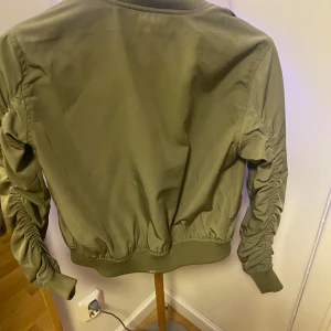 Olivgrön bomberjacka från FB Sister - Snygg olivgrön bomberjacka från FB Sister i storlek XS. Jackan har dragkedja framtill, ribbade muddar och coola rynkade detaljer längs ärmarna. Materialet är mjukt och lätt, perfekt för att ge din outfit en streetstyle-vibe.