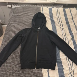 Jack & Jones Hoddie - Svart hoodie från Jack & Jones hel dragkedja framtill och två stora fickor. Tröjan har huva med snörning och långa ärmar. Materialet är mjukt och känns som bomull eller polyester, perfekt för chill dagar. Enkel och stilren design utan tryck eller mönster.