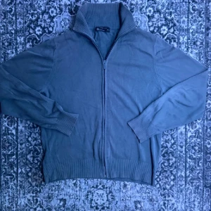 Grå kofta med dragkedja från Zara Man - Snygg grå kofta från Zara Man i storlek M. Koftan har en ribbad hög krage, långa ärmar och dragkedja framtill. Tillverkad i mjuk bomull och polyester, perfekt för att hålla stilen när det är lite kyligare ute.