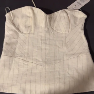 Vit randig korsettopp från Bershka - Säljer en snygg vit korsettopp från Bershka med tunna vertikala ränder och axelbandslös design. Toppen har markerade kupor och snörning i ryggen för perfekt passform. Materialet är mjukt och lätt, perfekt till sommarens outfits.