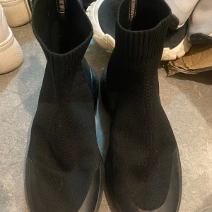 Svarta stickade boots med platt sula - Svarta boots med stickad ovandel och ribbad skaft som sitter tight runt ankeln. Skorna har en platt, grov sula och är helt svarta med en sportig och modern look. Perfekta för dig som gillar streetstyle och vill ha något bekvämt men ändå snyggt.