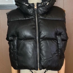 Snygg väst från Bershka - Tuff skinn imitations väst med huva från Bershka Västen är helt ny och oanvänd i perfekt skick! Det är en varm vadderad modell med glansigt innerfoder Västen har 2st fickor framtill, en bred silverfärgad dragkedja och den är ställbar nertill Längd: 47cm Bredd under ärm: 56x2cm Det är en storlek Large, men den passar bra till en Medium/38