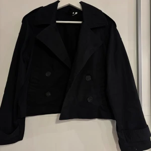 Svart skepparkavaj med knappar - Snygg svart skepparkavaj med dubbelknäppning och bred krage. Jackan är croppad och har långa ärmar med klassiska manschetter. Perfekt för dig som gillar stilrena och tidlösa plagg med en cool edge.