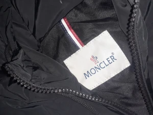 Svart vindjacka från Moncler - Svart vindjacka från Moncler med huva och dragkedja framtill. Jackan har Moncler-logga på ärmen och snygga detaljer med röd och vit rand på huvan. Tillverkad i lätt polyester, perfekt för blåsiga dagar. Fickor med knappstängning och justerbar huva.