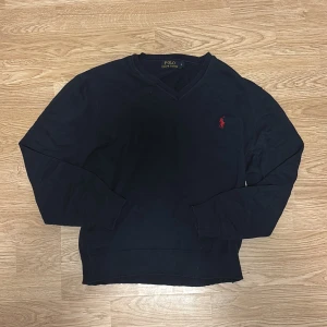 Marinblå v-ringad tröja Polo Ralph Lauren - Klassisk marinblå v-ringad tröja från Polo Ralph Lauren med röd broderad logga på bröstet. Tröjan är långärmad och har ribbade muddar vid ärmslut och nederkant. Perfekt för en stilren och avslappnad look.