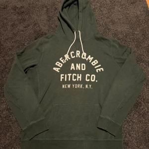 Mörkgrön hoodie Abercrombie & Fitch - Mörkgrön hoodie från Abercrombie & Fitch med vit broderad logga på bröstet. Klassisk känguruficka, huva med vita snören och långa ärmar. Perfekt för chill dagar och snygg streetstyle.