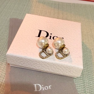 Dior örhängen med pärlor och strass - Supersnygga örhängen från Dior med dubbla vita pärlor och guldfärgade detaljer. Framsidan har en mindre pärla och baksidan en större, samt en ikonisk Dior-logga dekorerad med glittrande strass. Perfekt statement-piece för dig som vill sticka ut.