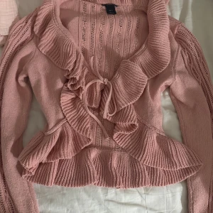 Rosa volangkofta med knyt från kaphal - Supersöt rosa stickad kofta från H&M med volangkant och knyt framtill. Koftan har långärmade ärmar med fina stickade detaljer och en croppad passform. 