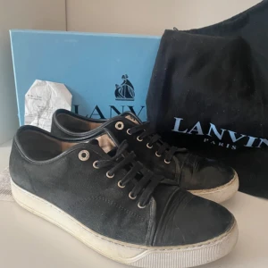 Lanvin skor - Svarta sneakers från Lanvin med rund tå och platt vit sula. Skorna har ovandel i skinn och mocka, svarta skosnören och metallfärgade öljetter. Klassisk och stilren design med diskreta detaljer, levereras med originalkartong och dustbag.