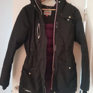 Svart parkajacka från McKinley, stl 36 - Svart parkajacka från McKinley med huva, stora fickor och dragkedja framtill. Jackan är fodrad med lila polyester och har justerbar midja. Perfekt för kyliga dagar 