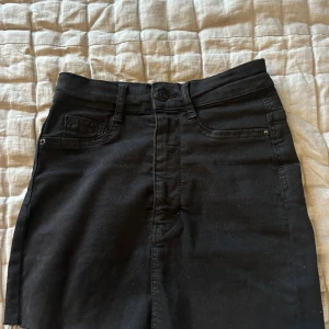 Svart jeanskjol med rå kant - Svart kort jeanskjol från Gina tricot. Klassiska fem fickor och rå, fransig nederkant. Kjolen har bälteshällor och knappstängning framtill. Perfekt för en cool och avslappnad stil.