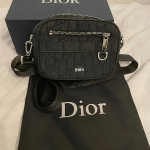 Svart Dior axelväska med monogram - Säljer en svart axelväska från Dior med ikoniskt monogrammönster och silverfärgade detaljer. Väskan har justerbar axelrem, dragkedja upptill och ett extra fack framtill med dragkedja. Insidan har kortfack och väskan är tillverkad i textil med detaljer i skinn.