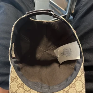 Gucci beige keps med GG-mönster - Säljer en snygg beige keps från Gucci med klassiskt GG-mönster över hela kepsen. Den har grönt och rött band på sidan och justerbar rem i brunt skinn baktill. Materialet är bomull och skinn, perfekt för dig som vill ha en lyxig streetstyle.